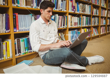 Male student typing on laptop while sitting on library floor with open textbook and notebook Male student typing on laptop while sitting on library floor with open textbook and notebook 129584887