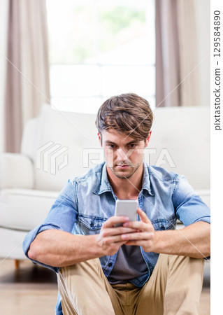 Man in mid-twenties sitting on wooden floor before white sofa in living room holding smartphone 129584890