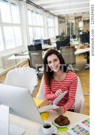 Female designer sitting at desk reviewing colorful folders in modern office with computer monitor 129584893