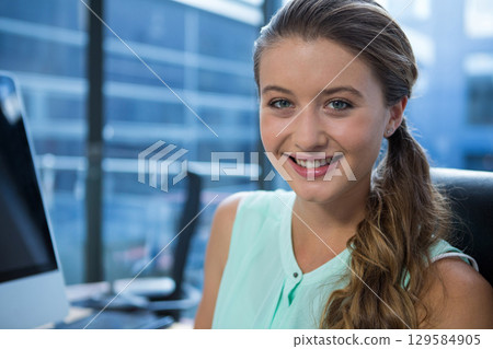 Female office worker wearing mint blouse smiling using computer by windows at office, copy space 129584905