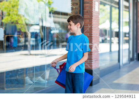 Boy standing outside retail store holding shopping bags, gazing at mannequins, copy space 129584911