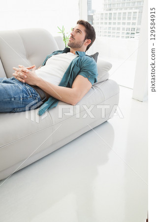 Man reclining on white leather sofa in living room with potted plant, skyline view, copy space 129584915