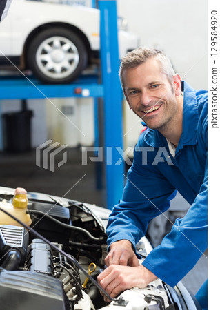 Mid adult man in coveralls leaning over open hood checking engine at repair shop, copy space 129584920