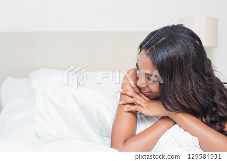 African American woman reclining on bed in white sheets, smiling, gazing right by lamp, copy space 129584931