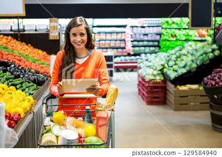 Woman holding tablet above shopping cart loaded with baguette cereal and produce in produce aisle 129584940