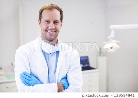 Male dentist standing crossing arms in dental exam room showing exam light laptop cabinetry 129584941
