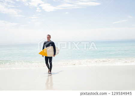 Male surfer walking barefoot on sandy beach by turquoise sea carrying surfboard with yellow nose 129584945