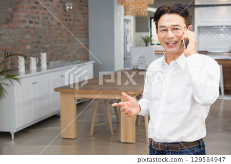 Senior Asian man standing near wooden dining table in modern kitchen holding smartphone, copy space 129584947
