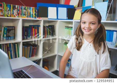 Girl wearing white blouse sitting at desk in school library using laptop near bookshelf, copy space 129584951