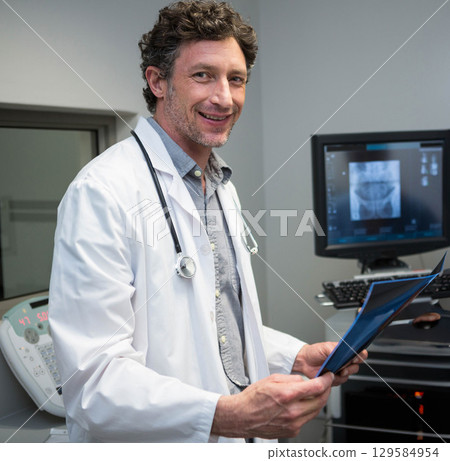 Male physician with stethoscope standing in exam room holding blue-edged radiograph beside console 129584954