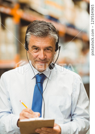 Mature man wearing white shirt and blue tie using headset and checking clipboard in warehouse Mature man wearing white shirt and blue tie using headset and checking clipboard in warehouse 129584965