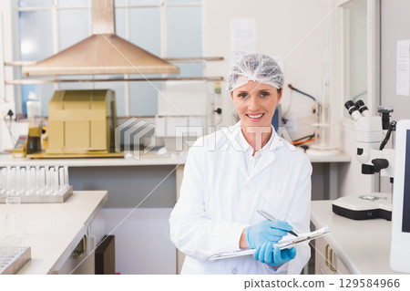 Woman wearing lab coat and gloves standing at lab bench with microscope and clipboard, copy space 129584966