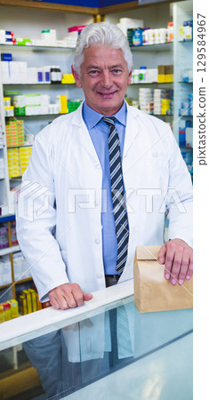 Male pharmacist in lab coat placing brown paper bag on glass-topped counter at pharmacy 129584967