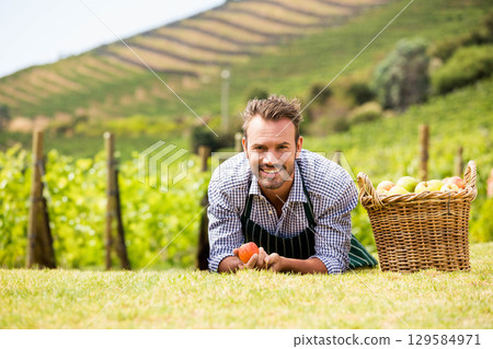 Man in striped apron lying on orchard grass holding ripe apple and smiling beside wicker basket 129584971