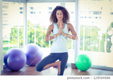 Woman practicing yoga tree pose on wooden floor by windows with purple and green exercise balls 129584986