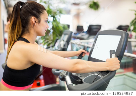 Female fitness enthusiast gripping elliptical handles monitoring digital console at gym, copy space 129584987