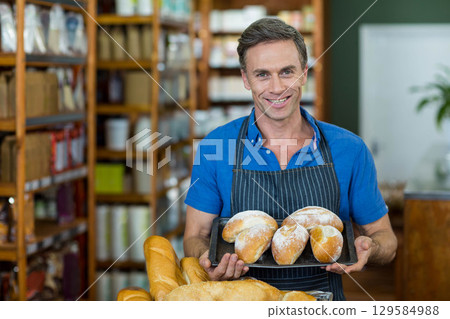 Mature male baker in dark apron presenting tray of four flour-dusted loaves at bakery counter 129584988