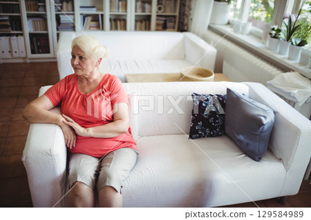 Senior woman sitting on sofa in living room wearing coral top beside built-in bookshelf 129584989