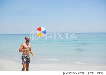 Shirtless man in floral swim trunks tossing multicolored beach ball on sand shoreline, copy space Shirtless man in floral swim trunks tossing multicolored beach ball on sand shoreline, copy space 129584991