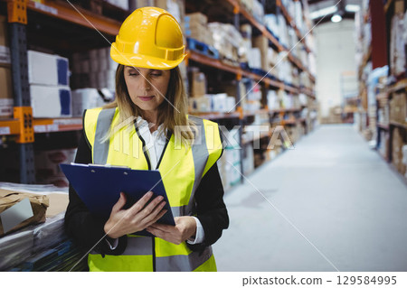 Female supervisor wearing helmet and vest standing beside pallet racks reviewing stock on clipboard Female supervisor wearing helmet and vest standing beside pallet racks reviewing stock on clipboard 129584995