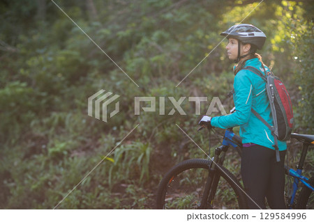 Female cyclist standing by mountain bike on wooded trail wearing helmet gloves backpack, copy space 129584996