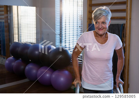 Senior woman balancing on parallel bars at gym with stability balls, mirror, blinds, copy space 129585005