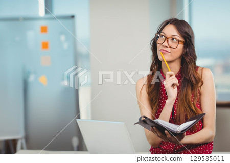 Woman sitting at desk in modern office holding notebook and pencil before laptop, copy space Woman sitting at desk in modern office holding notebook and pencil before laptop, copy space 129585011