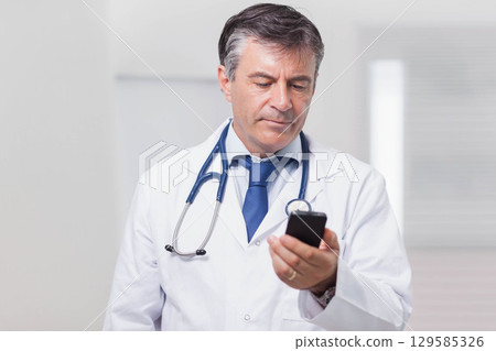 Mature male doctor standing in clinic corridor wearing lab coat, stethoscope while using smartphone 129585326