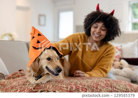 Woman wearing sweater petting golden retriever wearing jack-o-lantern hat on throw in living room 129585379