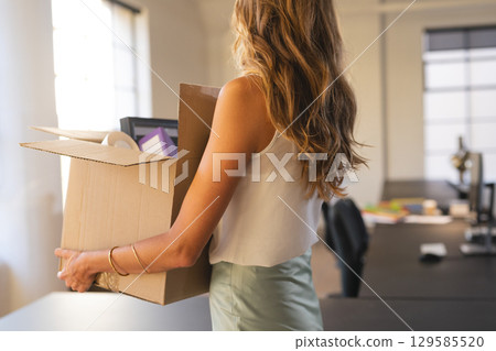 Mid adult woman holding box filled with documents and purple binder in open-plan office, copy space 129585520