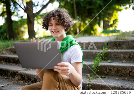 Curly-haired teen sitting on the steps in the park and having a video call Curly-haired teen sitting on the steps in the park and having a video call 129585639