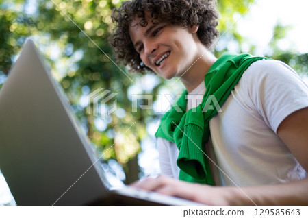 Curly-haired teen sitting on the steps in the park and having a video call Curly-haired teen sitting on the steps in the park and having a video call 129585643