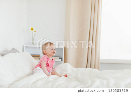 Baby girl sitting upright on bed in bedroom beside books, vase of yellow flowers, copy space 129585719