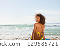African American woman in bikini sitting on towel at beach glancing back at waves, copy space 129585721