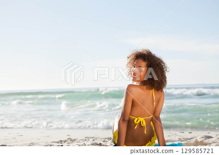 African American woman in bikini sitting on towel at beach glancing back at waves, copy space African American woman in bikini sitting on towel at beach glancing back at waves, copy space 129585721