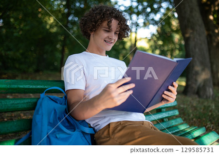 Curly-haired teenager sitting on the bench in the park and reading a book Curly-haired teenager sitting on the bench in the park and reading a book 129585731