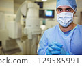 Male surgeon wearing blue gown, mask, gloves beside C-arm and monitors in imaging room, copy space 129585991