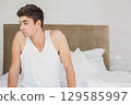 Man in his twenties sitting on bed edge in softly lit bedroom with small bedside lamp 129585997