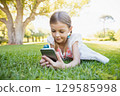 School-age girl lying on green lawn in sunlit park wearing red embroidered dress holding smartphone 129585998