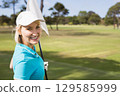 Senior woman wearing turquoise shirt holding flagstick on golf green with fairway view, copy space 129585999