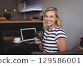 Woman sitting at coffee shop table holding smartphone, typing on laptop with coffee cup, copy space 129586001