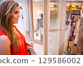Female shopper standing at mall store window holding polka-dot bag gazing at mannequins, copy space 129586002