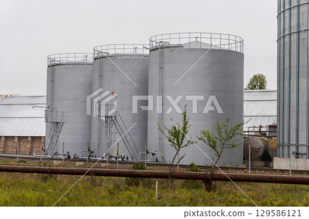 Tall grey silos/tanks in grassy field. Railway tracks, pipeline in front. Overcast 129586121
