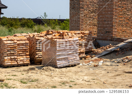 Stacks of clay bricks sit on pallets near a half-built brick wall at a construction site on a bright day, prepared for building a house Stacks of clay bricks sit on pallets near a half-built brick wall at a construction site on a bright day, prepared for building a house 129586153