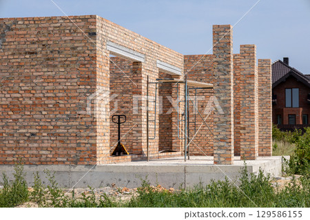 A brick house under construction has doorways and supporting pillars. Scaffolding and a hand truck are present A brick house under construction has doorways and supporting pillars. Scaffolding and a hand truck are present 129586155