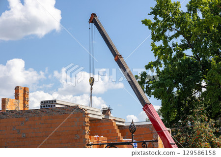 A red and black crane lifts precast concrete slabs to the top of a brick building under construction, set against a blue sky with white clouds 129586158