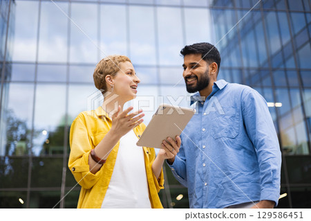Two colleagues discuss something on a tablet in front of a modern building. Two colleagues discuss something on a tablet in front of a modern building. 129586451