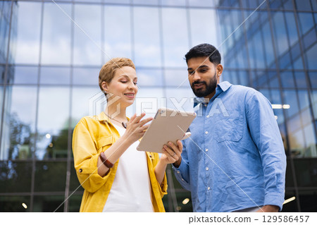 Two professionals review information on a tablet in front of a modern glass building, discussing a project. 129586457