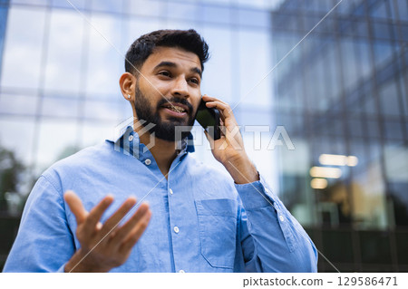 An Indian man talks on the phone outside a modern glass building, gesturing with his hand. 129586471