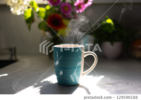 Warm Cup of Coffee on a Sunny Kitchen Counter With Flowers in the Background Warm Cup of Coffee on a Sunny Kitchen Counter With Flowers in the Background 129590188